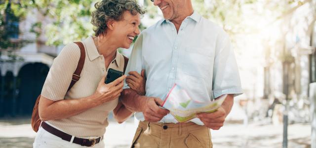 Elderly man and woman laughing. The man is holding a map, and the woman is wearing a backpack. They look like they are travelling.